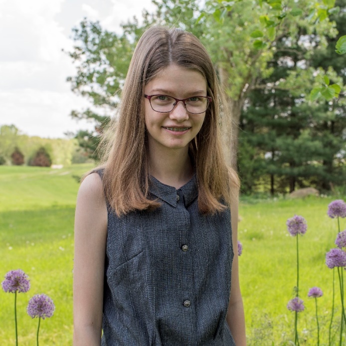 Phoebe Adair Harris, Chelsea, Michigan Young woman with blonde hair and gray sleeveless top stands among flowers with grass and trees behind her