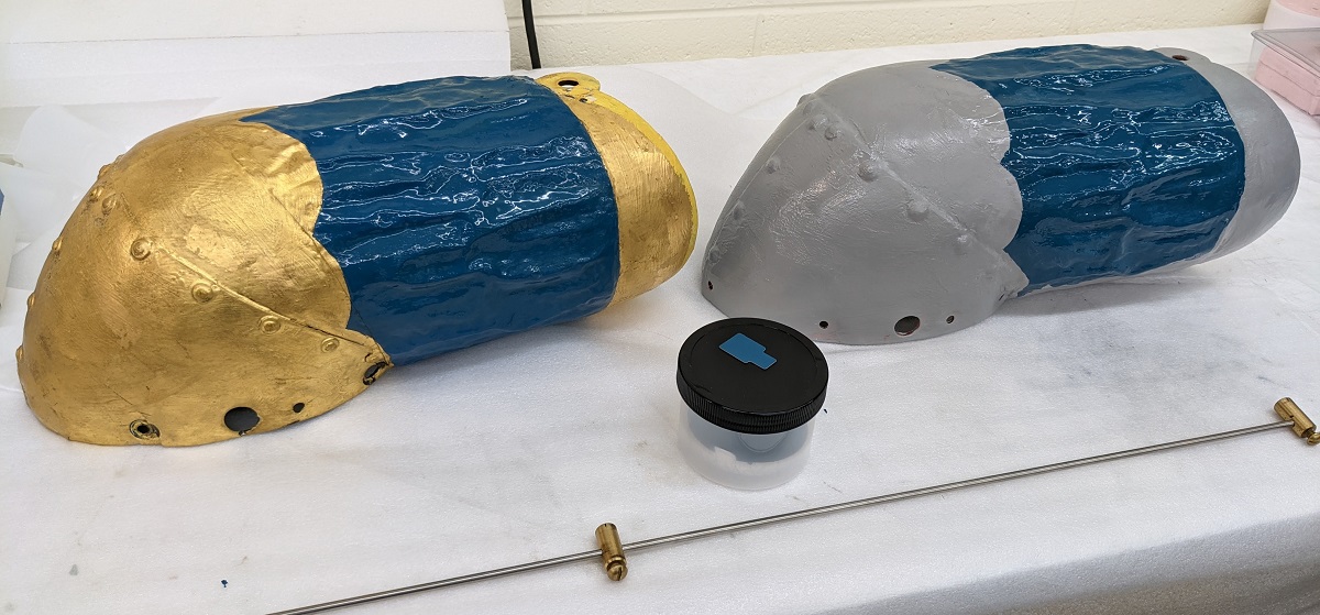 Original arm, left; 3D-printed arm, right; and Munsell color sample in the middle Two identical tubular shapes next to each other, one gold and blue and one gray and blue, with small jar between them