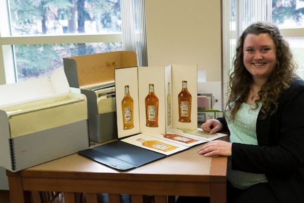 Project Curator Samantha Johnson with Items from the H.J. Heinz Company Collection in the Benson Ford Research Center. Woman sitting at table with boxes and images both standing up and lying flat