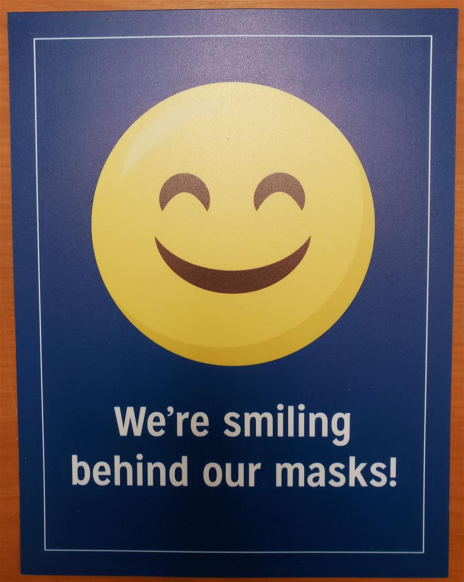"We're smiling behind our masks" sign from Henry Ford Health System Yellow "smiley face" emoji; also contains text