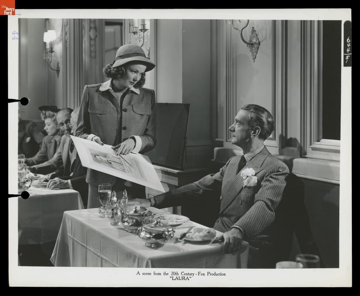 Black-and-white photo of a woman in a suit and hat standing at a table in a restaurant showing a poster to a man seated at a table