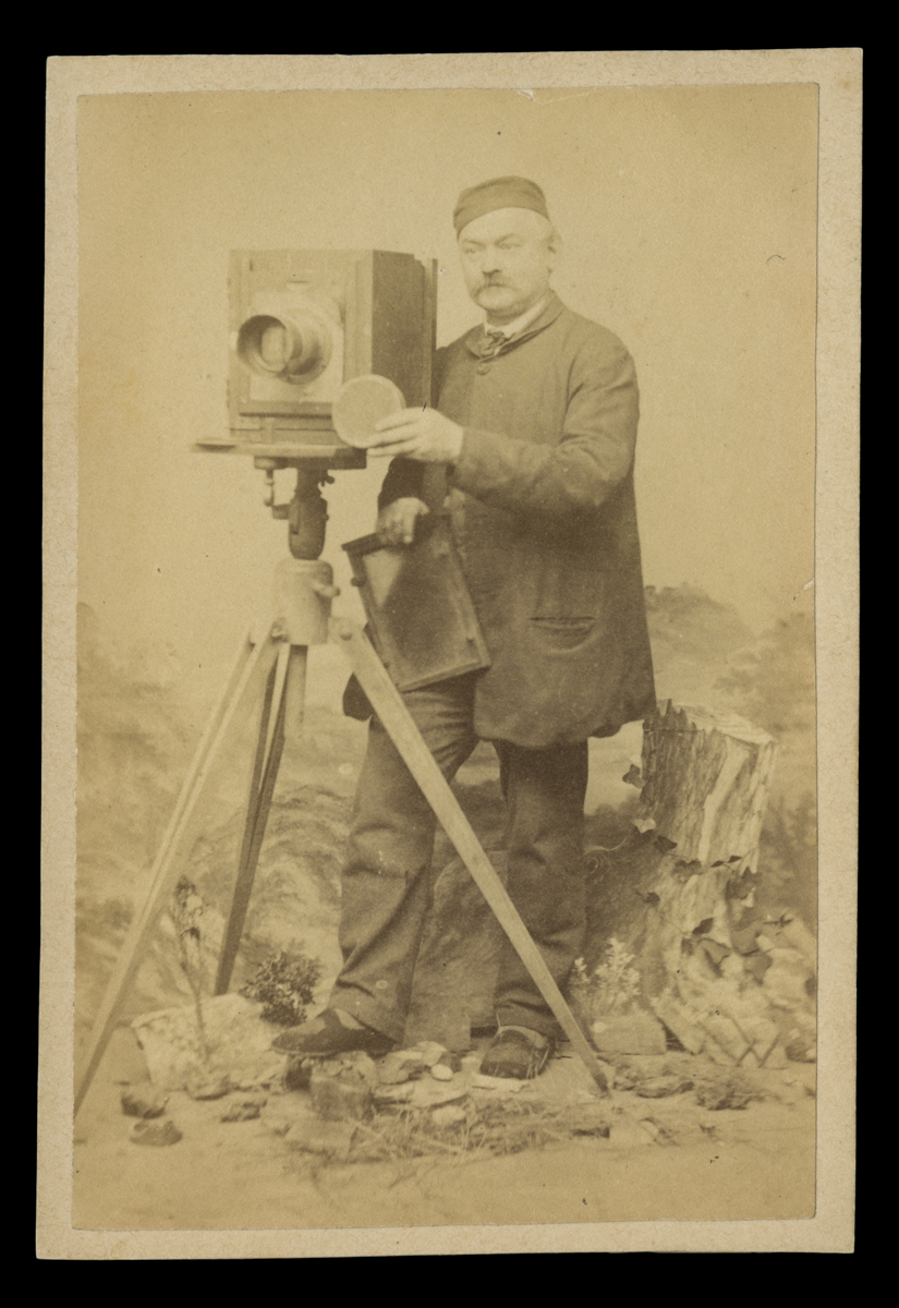 Man in smock and cap standing behind camera on tripod