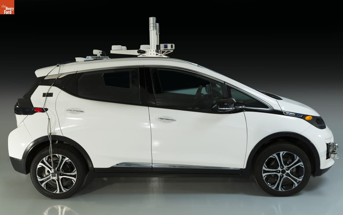 2016 General Motors First-Generation Self-Driving Test Vehicle Side view of compact white car with equipment on top and wires dangling down side