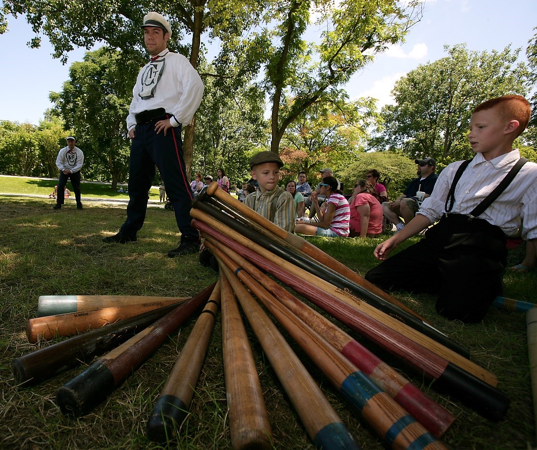 Historic Base Ball in Greenfield Village A variety of wooden bats is piled intricately in the foreground, with bat boys and historic base ball players in costume nearby, and a crowd watching the game sits behind them