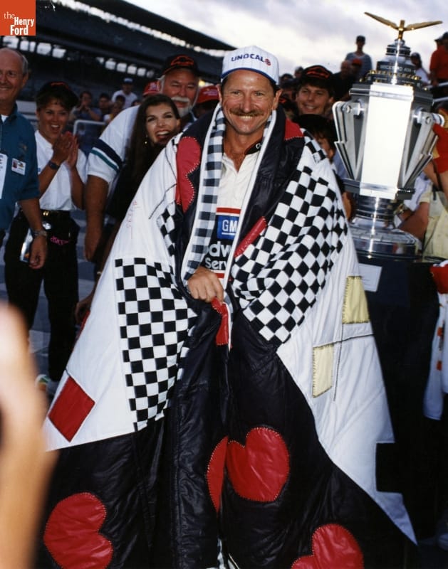 Dale Earnhardt with a Quilt Made by Jeanetta Holder, after the 1995 Brickyard 400 Race at the Indianapolis Motor Speedway Man wrapped in quilt wearing baseball cap stands among other people with a large trophy and grandstands in the background