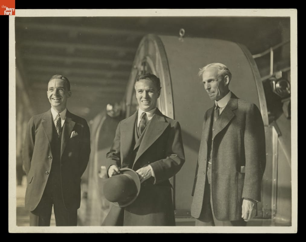 Edsel Ford, Charlie Chaplin, and Henry Ford Touring the Ford Motor Company Highland Park Plant, October 1923 Three men in suits, one in middle holding hat, pose for a photo in front of large equipment or machinery