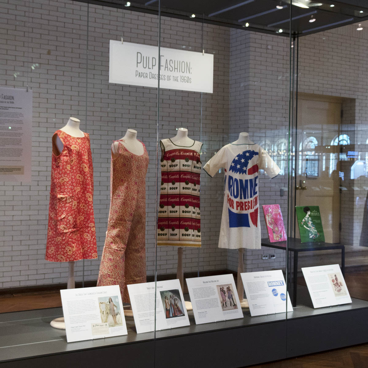 “What We Wore” case in Henry Ford Museum, featuring “Pulp Fashion” paper dresses exhibit Glass case with four dress forms, each containing a garment, labels in front and additional artifacts on a low table