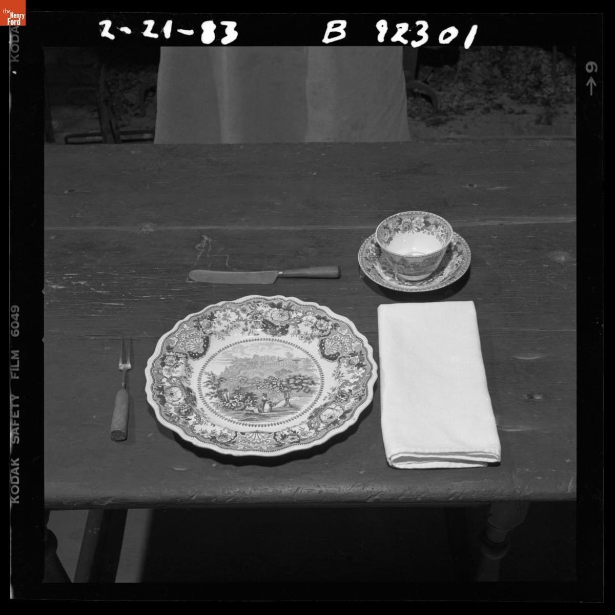 Black-and-white photo of table setting with plate, fork, knife, napkin, cup, and saucer on wooden table