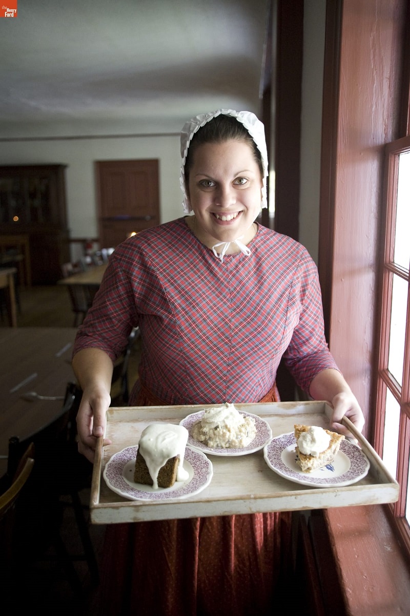 Woman in pink plaid dress and white bonnet smiles and holds tray of pastries