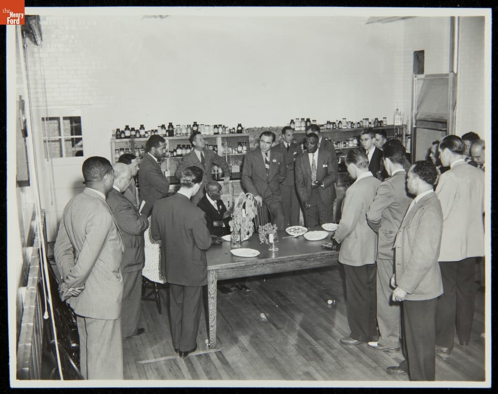 Group of people stands around a large table in a room with shelves filled with many small bottles and jars