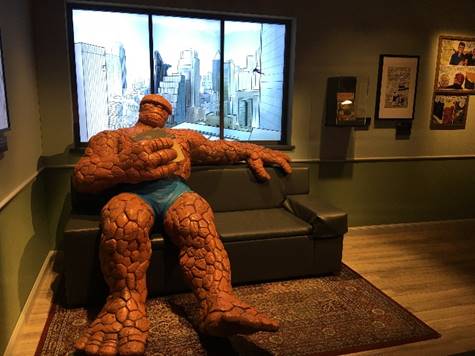 Oversize mannequin with orange reptilian skin sitting on couch with illuminated drawing of city skyline behind him; additional cases and framed drawings on wall to right