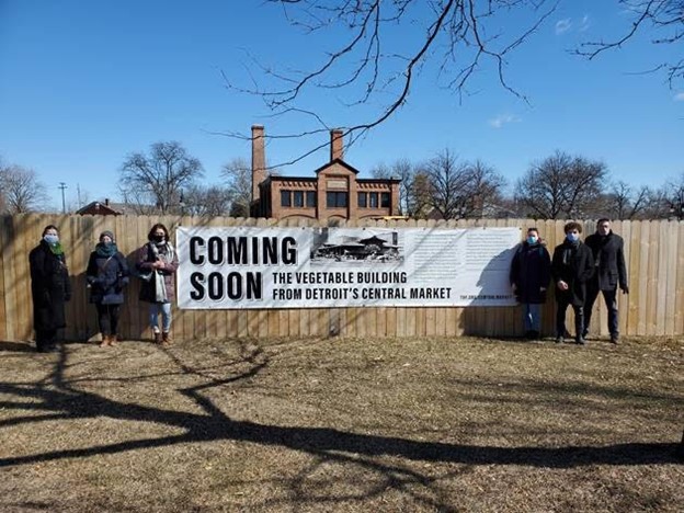 Six people pose in front of a wooden fence on either side of a sign reading "Coming Soon: The Vegetable Building from Detroit's Central Market"