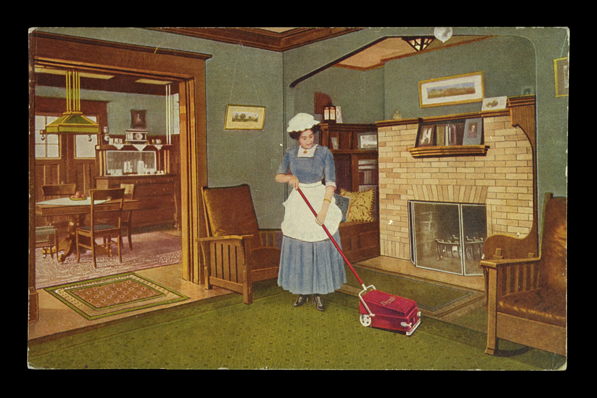 Woman in blue dress with white apron and cap using a sweeper vacuum on a living room carpet