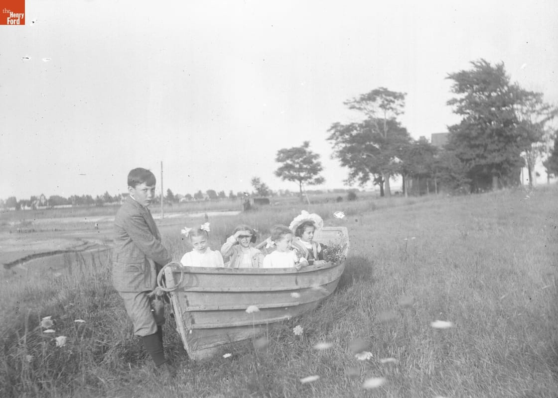 Crookhaven, Children in Boat, 1890-1915 Young boy pulls at the front of a wooden rowboat in a grassy field, while four girls sit inside