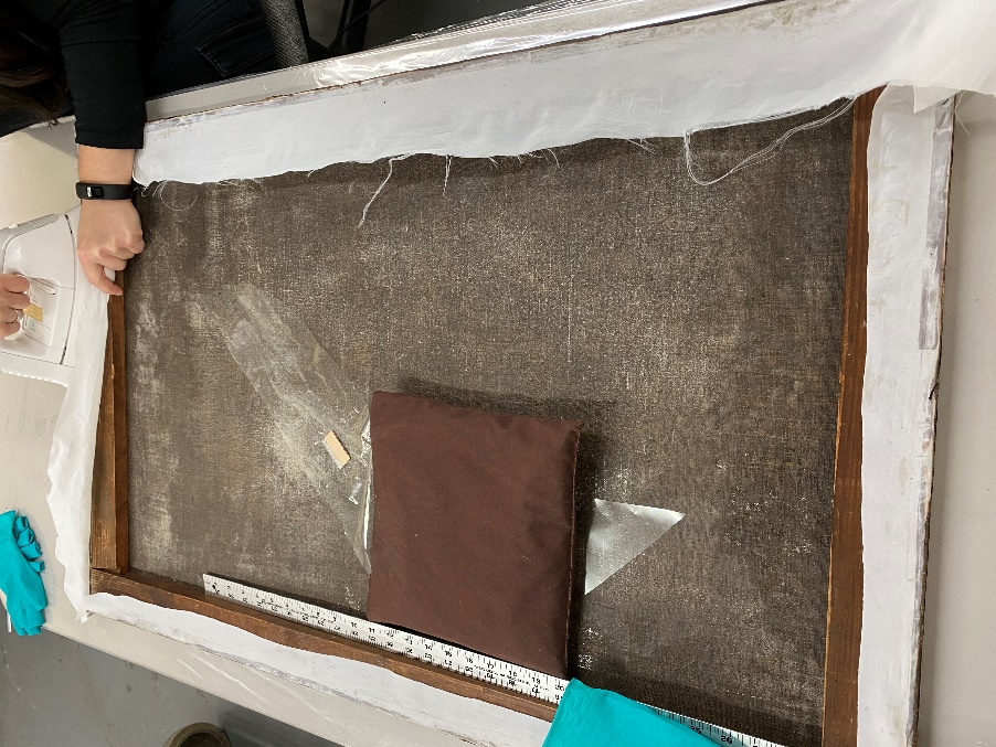 Canvas is re-stretched and extra sail cloth removed A person's hands work at top of painting laid facing down on flat table