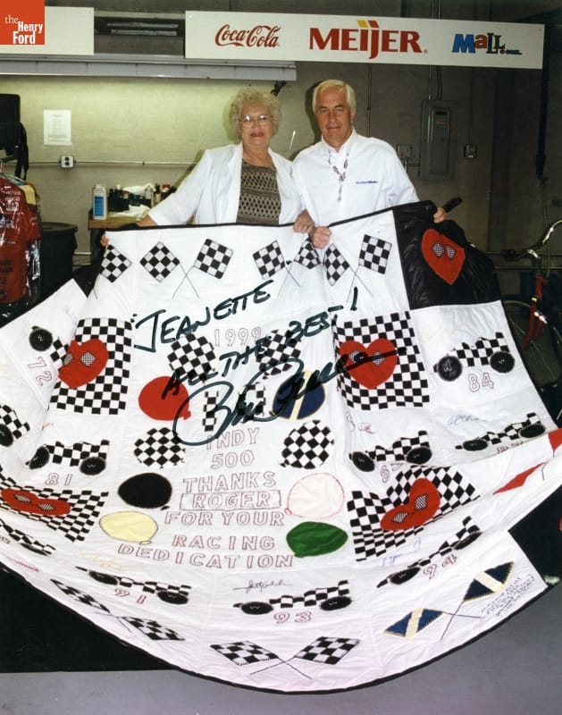 Woman with blonde hair and glasses and man with white hair in a white dress shirt hold a quilt; text inscription and signature on photograph