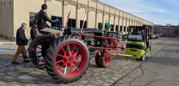Moving the tractor into the museum Small vehicle towing a large gray tractor with red wheels, with a man sitting on the tractor and a couple others walking next to it