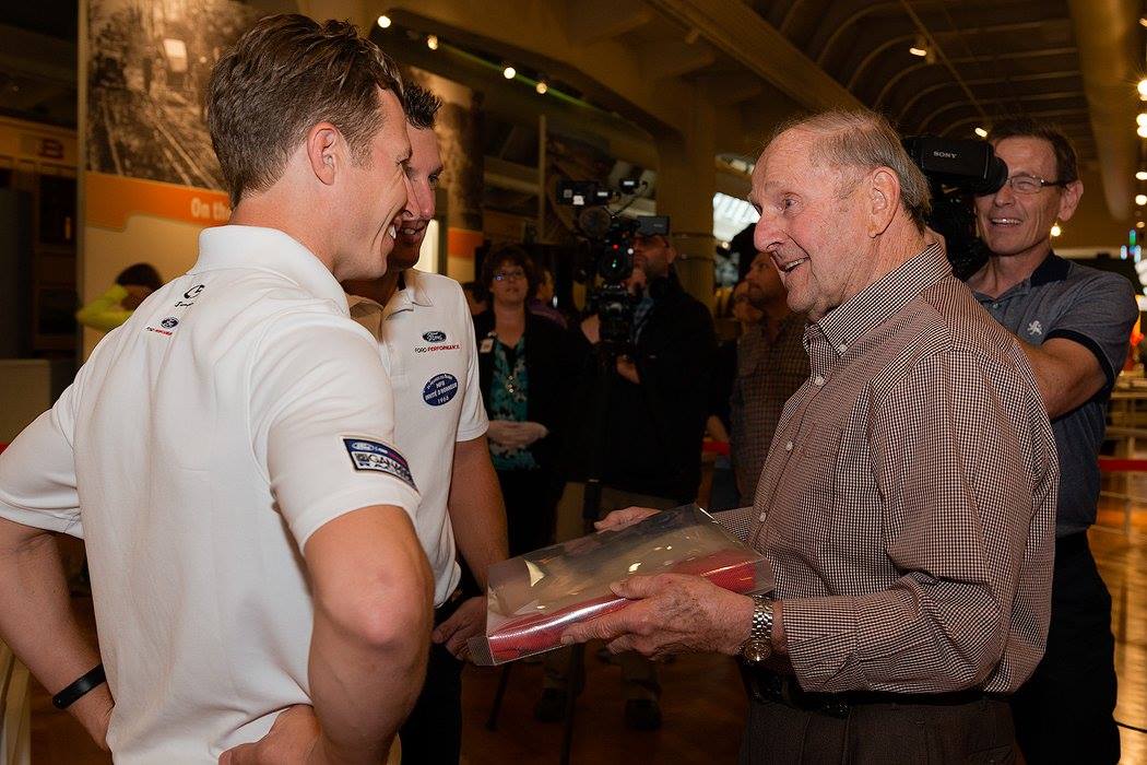 Mose Nowland lending Ford Performance drivers Ryan Briscoe and Joey Hand his souvenir French flag Smiling man hands a package to two other men, while people look on and camerapeople film