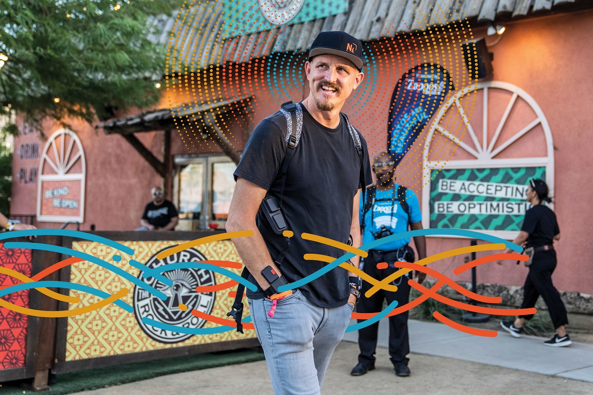 Mick Ebeling, founder of Not Impossible Labs Man in black t-shirt, jeans, baseball cap, and backpack stands in a plaza with a few other people and red building behind him