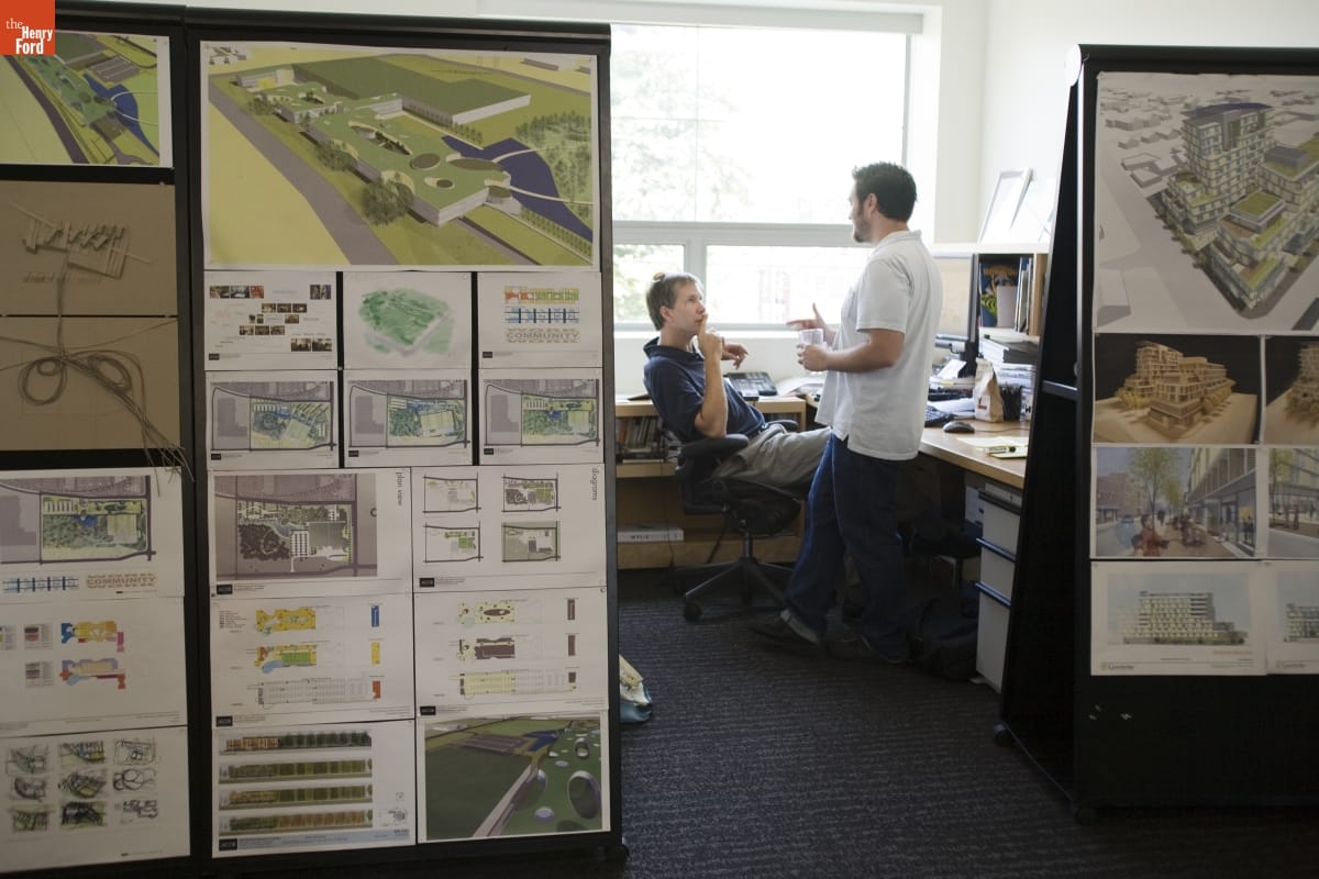 Oral History Interview with William McDonough, Sustainable Design Architect, September 2008--Photographs--Digital Images Two people, one seated and one standing, talk in a sunny office cubicle; drawings and renderings of buildings hang on outside walls