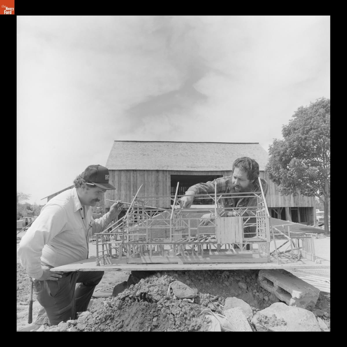 Two men look closely at a model of an unfinished building balanced on a pile of dirt with a wooden barn behind them
