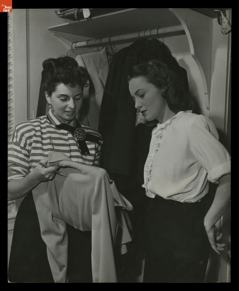 Black-and-white photo of two women standing in front of a closet with hanging clothing, looking at a blouse or jacket one of them is holding