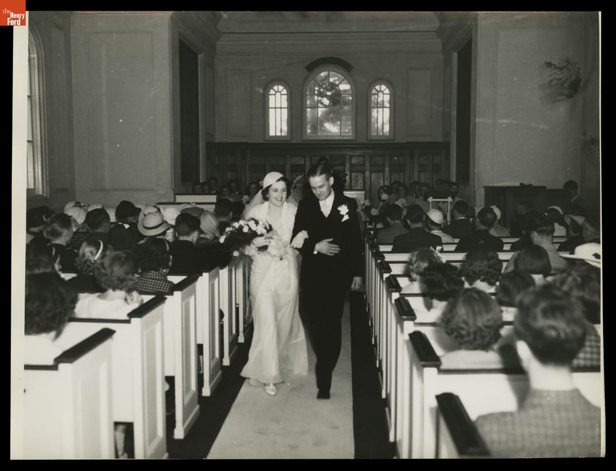 Bride and groom, arm in arm, walking down a church aisle past pews full of people sitting facing away from the camera