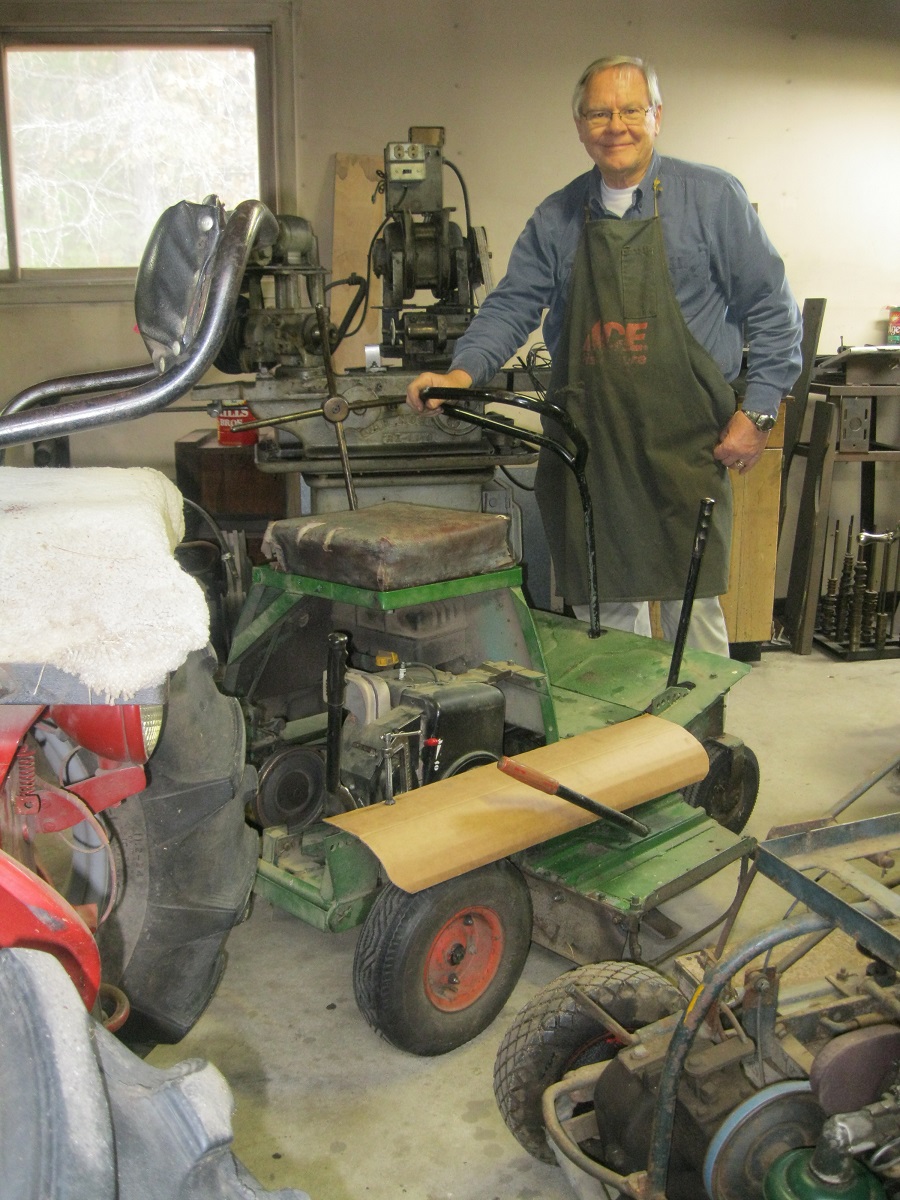 Curtis Swanson poses with one of his father’s prototype lawn mowers in November 2018 Man in denim shirt and shop apron stands in workshop next to machinery