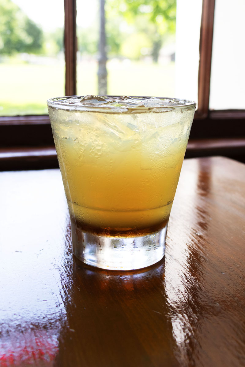 Eagle Tavern's Maple Bourbon Sour Honey-colored drink in clear glass with ice, sitting on wooden table in front of window