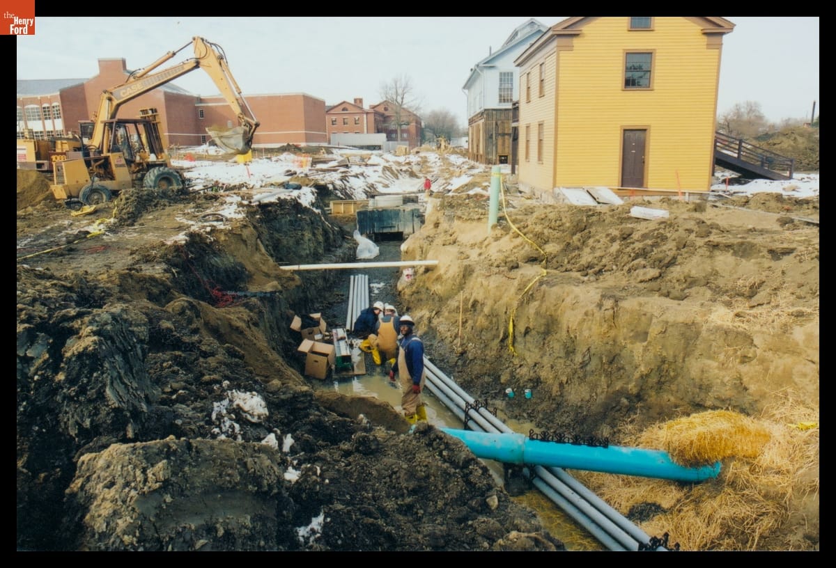 Three men with pipes in muddy trench; construction equipment and buildings nearby