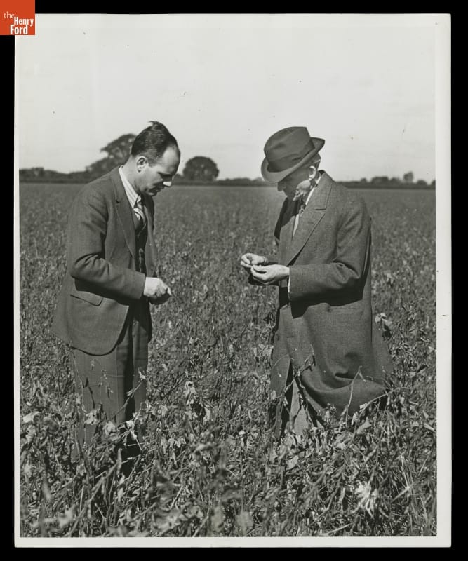 Two men in suits, one wearing a hat, look at something in a field