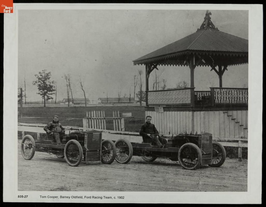 Tom Cooper and Barney Oldfield Seated in Race Cars, circa 1902 Two men pose at the wheel of two very minimal open early race cars on a track next to a covered pavilion