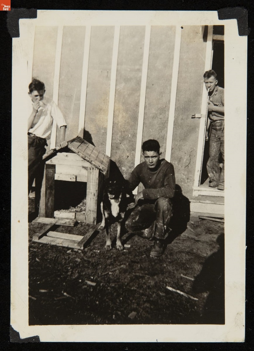 Man kneels with dog next to doghouse; other men stand nearby