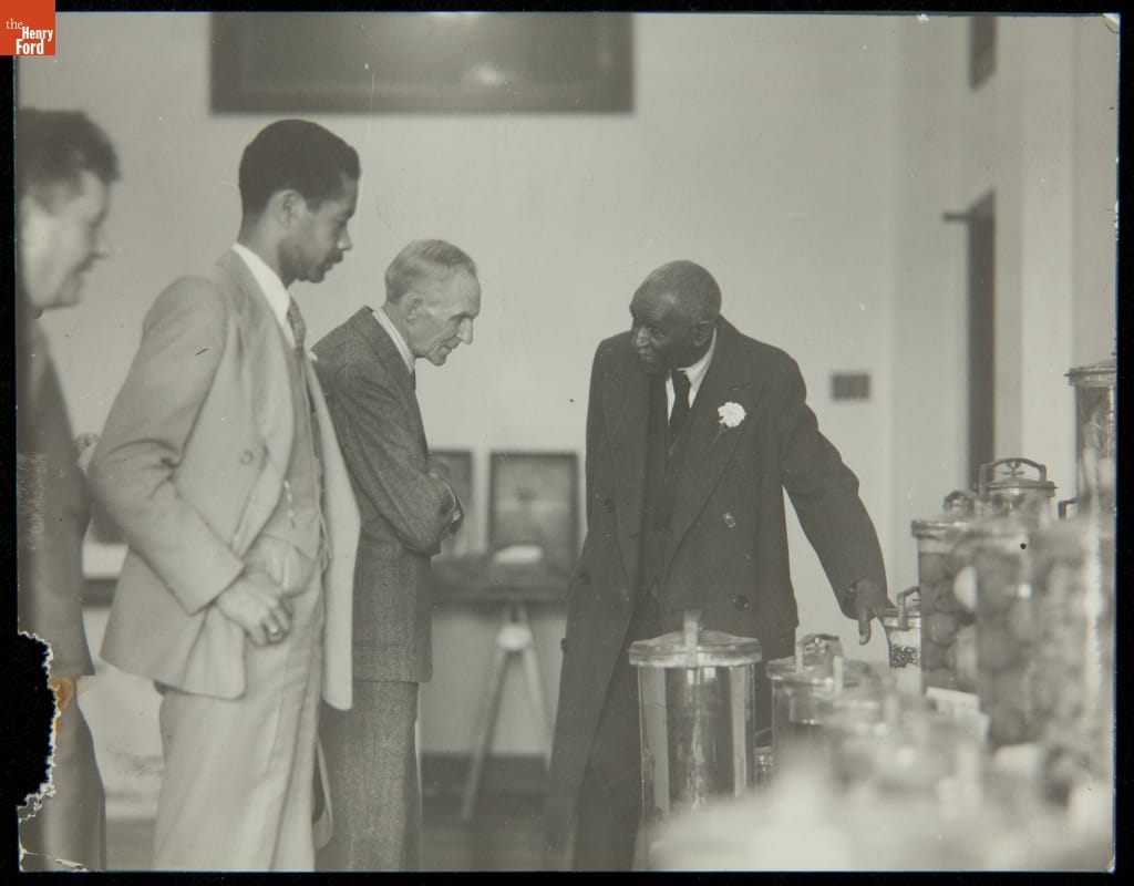 Frank Campsall, Austin Curtis, Henry Ford, and George Washington Carver at Tuskegee Institute, March 1938 Man gestures to a table covered in glass jars while other men look on