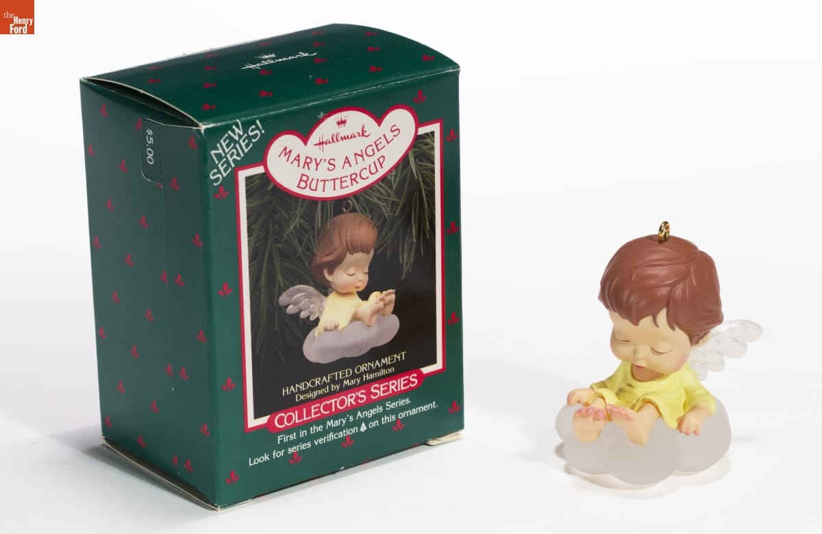 Hallmark "Mary's Angels Series: Buttercup" Christmas Ornament, 1988 Ornament with large-headed , brown-haired child/angel wearing a yellow dress with transparent wings sitting on transparent cloud; green box with image of ornament sits next to it