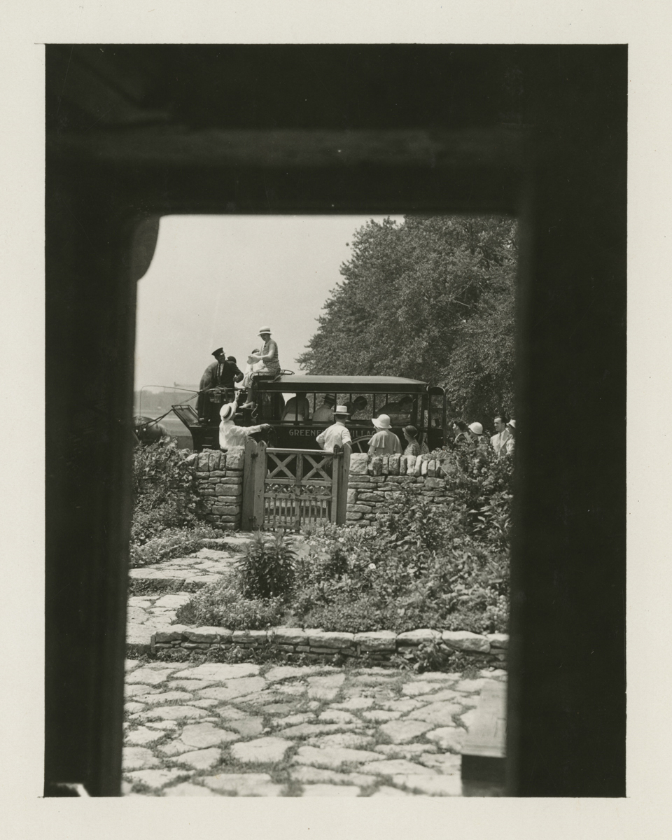 Photo taken from inside a window of people dismounting from an omnibus outside a garden with a gate