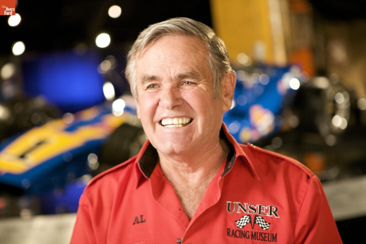 Oral History Interview with Al Unser Sr., January 6, 2009 / Photographed by Michelle Andonian Man in red shirt with text and checkered flag logo smiles at camera; out-of-focus race car in background