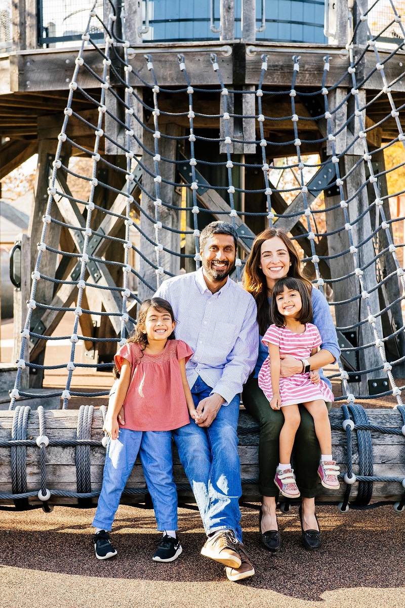 The Henry Ford members Meera, Sri, Maya, and Sonia Man, woman, and two young girls sit and stand on a log in front of netting attached to a playground structure
