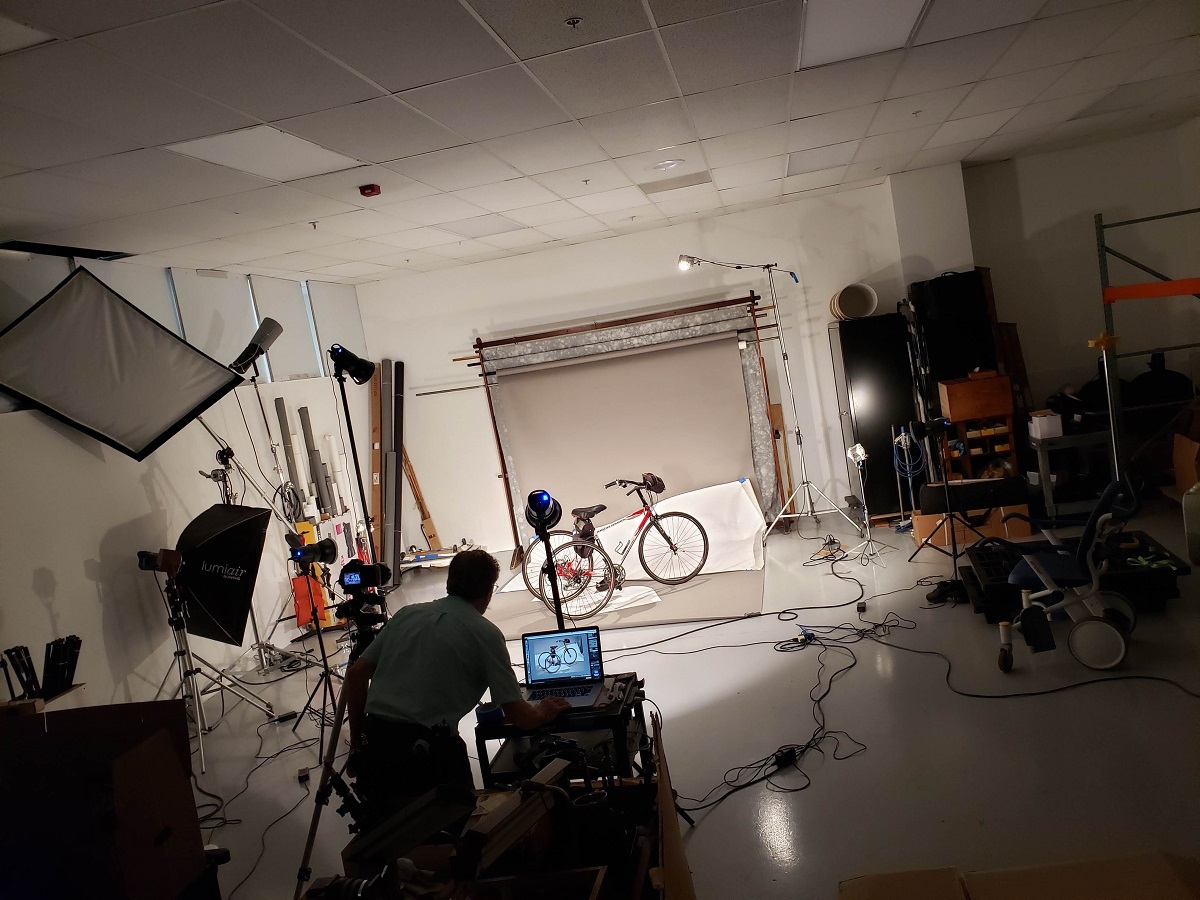 Man stands at a laptop on a cart in a photo studio with a tricycle on the set
