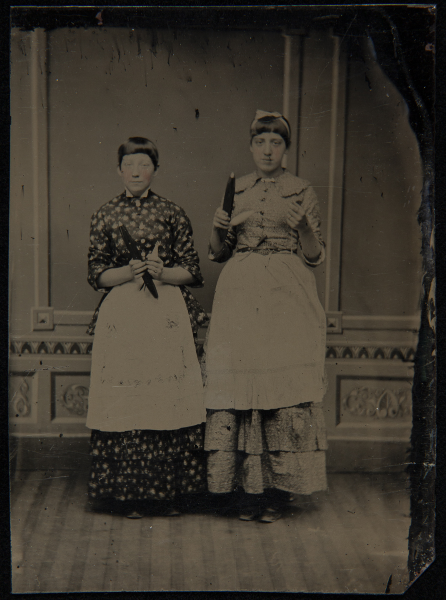 Two women with short hair wearing dresses and aprons holding spindles