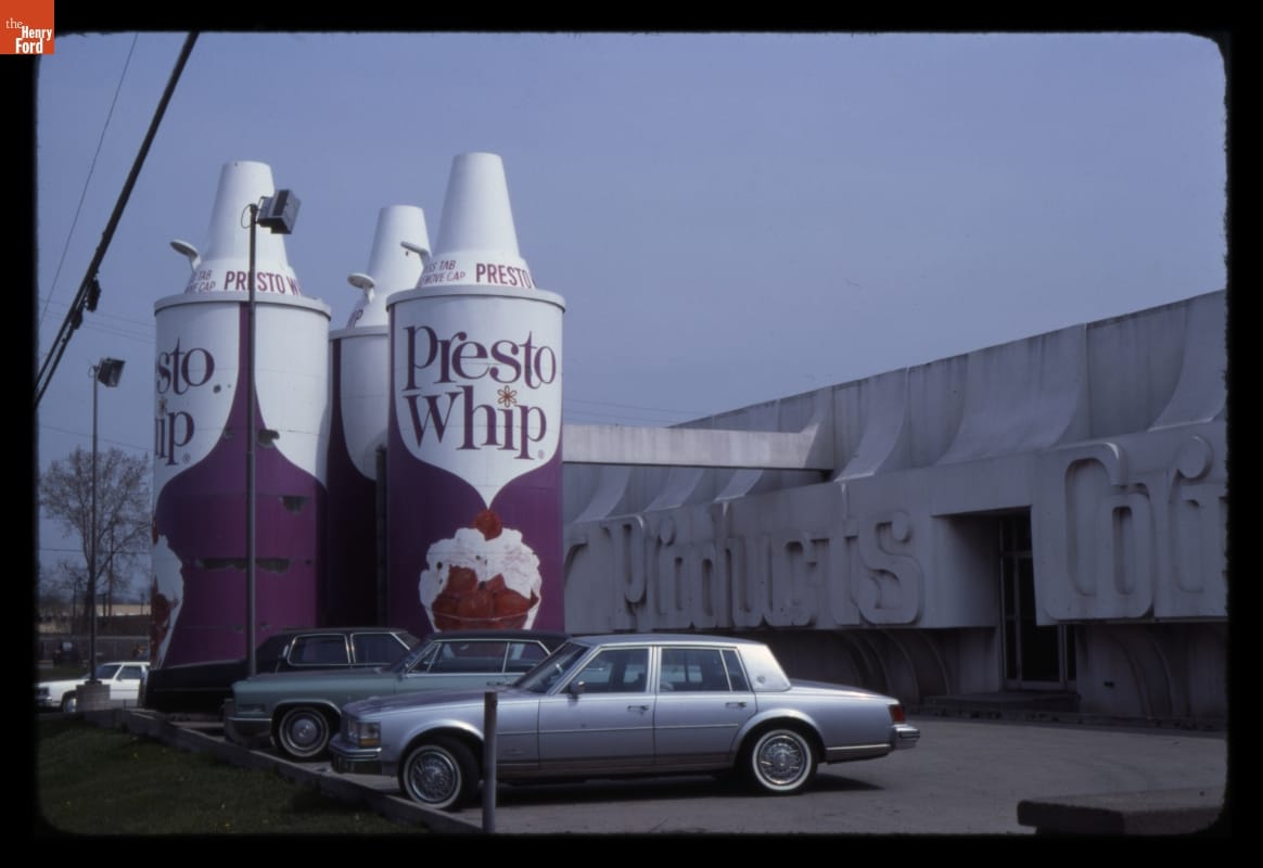 Building, parking lot, and something formed to look like three giantic cans of "Presto Whip" canned whipped cream