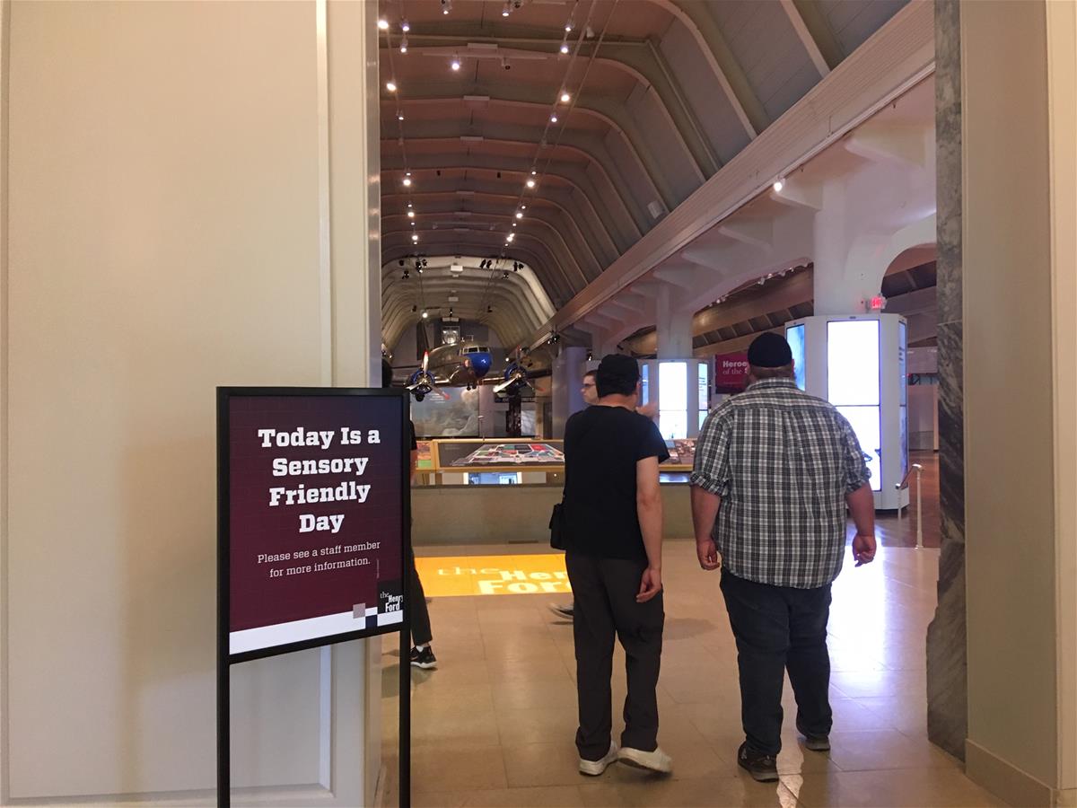 Sensory-friendly event sign at the entrance to Henry Ford Museum Two people walk past a sign with text into a large high room with an airplane hanging from the ceiling