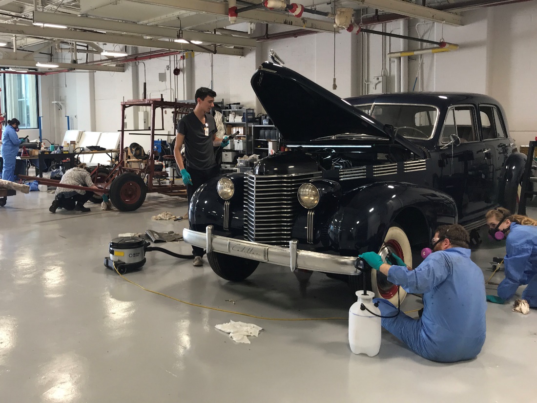 Our conservation team (pre-pandemic) cleans oversized artifacts A number of people, some wearing jumpsuits and respirators, work on cars and other equipment in a large room