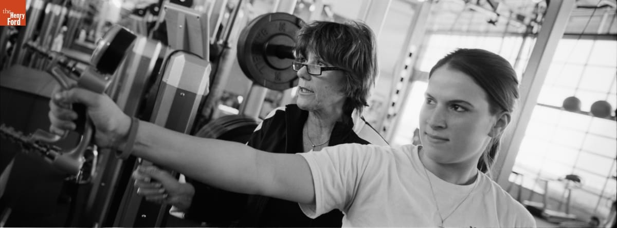 Lyn St. James Instructing a Student, 2008 Two women, one holding a piece of equipment in her hand, stand in a gym looking leftward