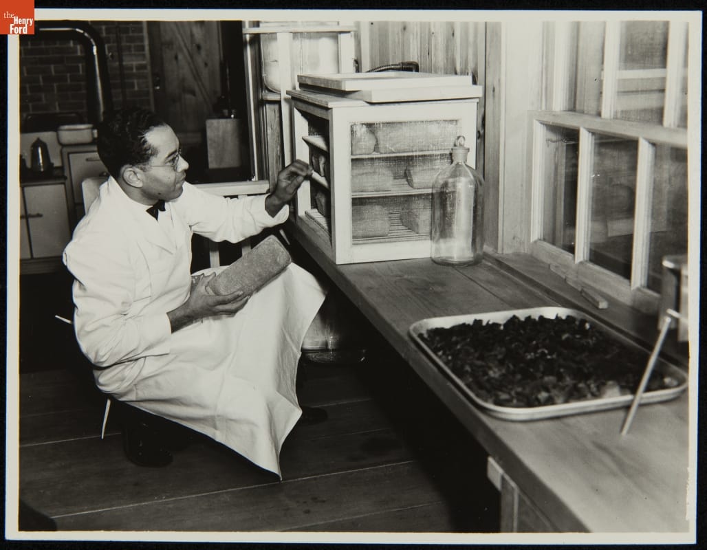 Man in white apron kneels with loaf of bread in his hand before small cabinet filled with loaves of bread