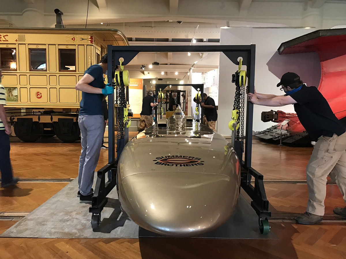 A forklift is attached to the rear gantry and used to tow Goldenrod into position over railroad tracks covered with steel plates. Men guide a golden car suspended from crane-like structures past railcars and streetcars