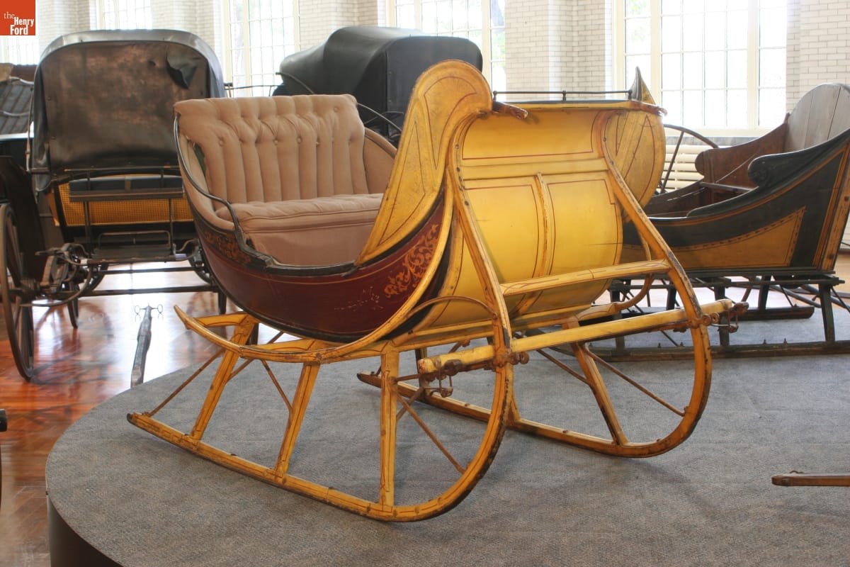 Albany Cutter, circa 1865 Yellow and maroon sleigh with beige upholstered seat, displayed in a room with other vehicles