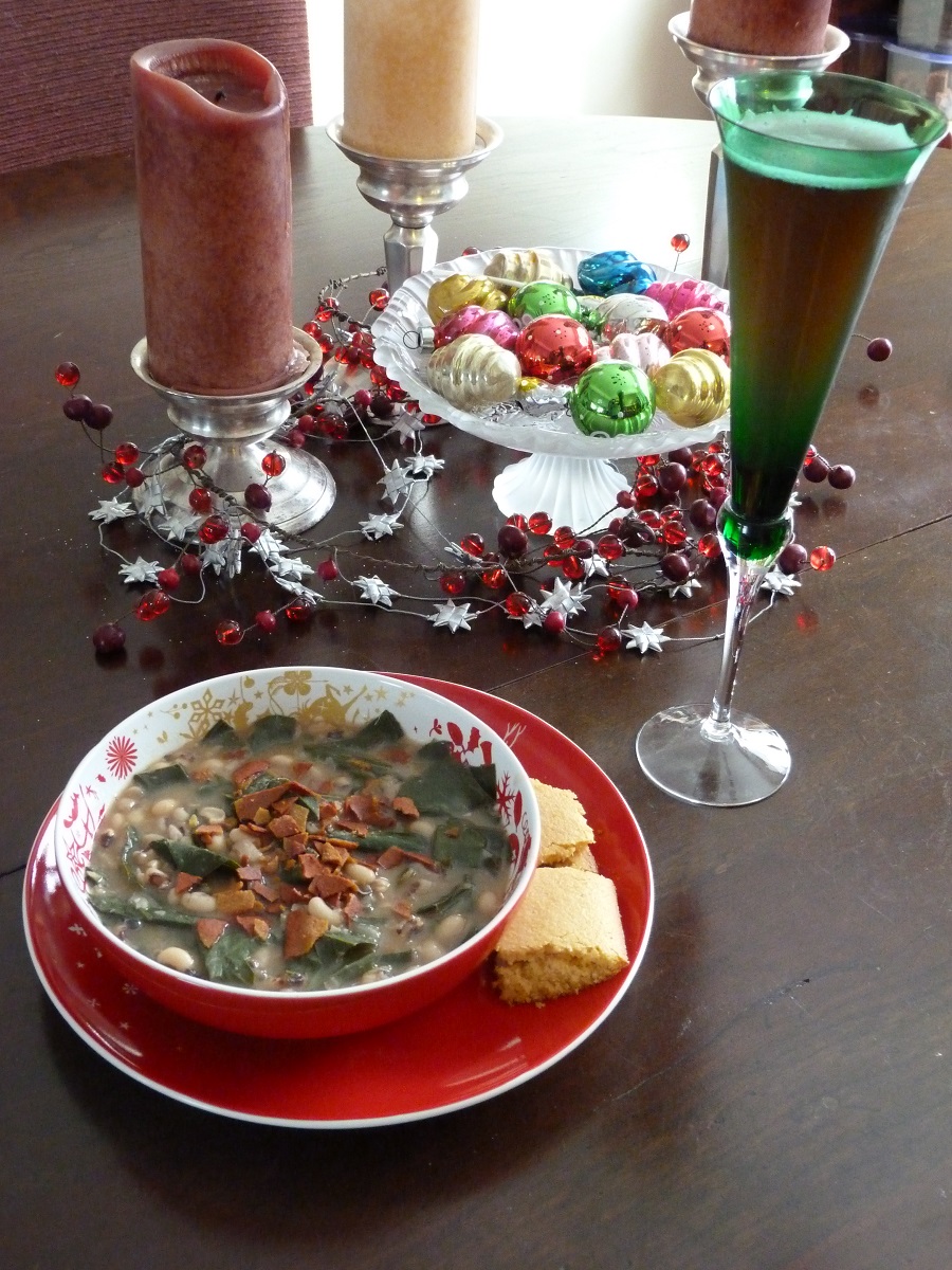 A vegetarian version of hoppin’ john Red dishware containing a soup with greens and veggie bacon, with cornbread on the side, a flute filled with liquid, and a holiday centerpiece with candles, ornaments, and garland