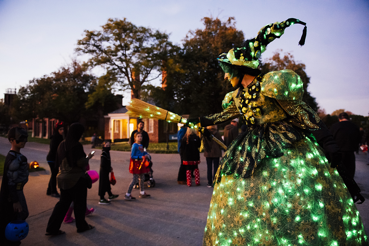 Woman in witch's costume decorated with green lights holds out a broom as children walk by