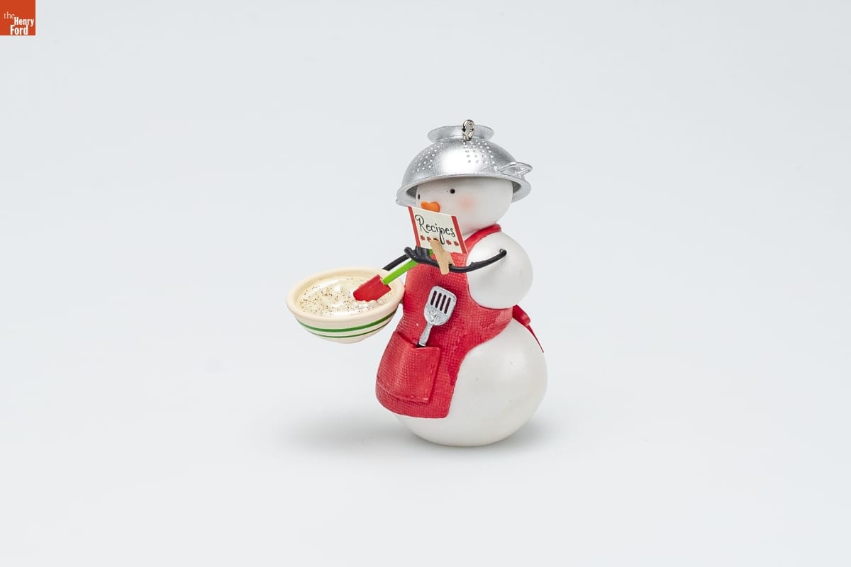 Hallmark "Snow Much Fun to Cook!" Christmas Ornament, 2009 Ornament of a snowman wearing a red apron with a spatula in the pocket and a colander on its head, holding a bowl of dough in one hand and a card labeled "Recipes" in the other